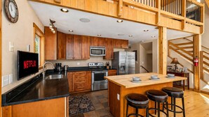 A modern kitchen with wooden cabinets, stainless steel appliances, a central island with stools, and a visible Netflix sign.