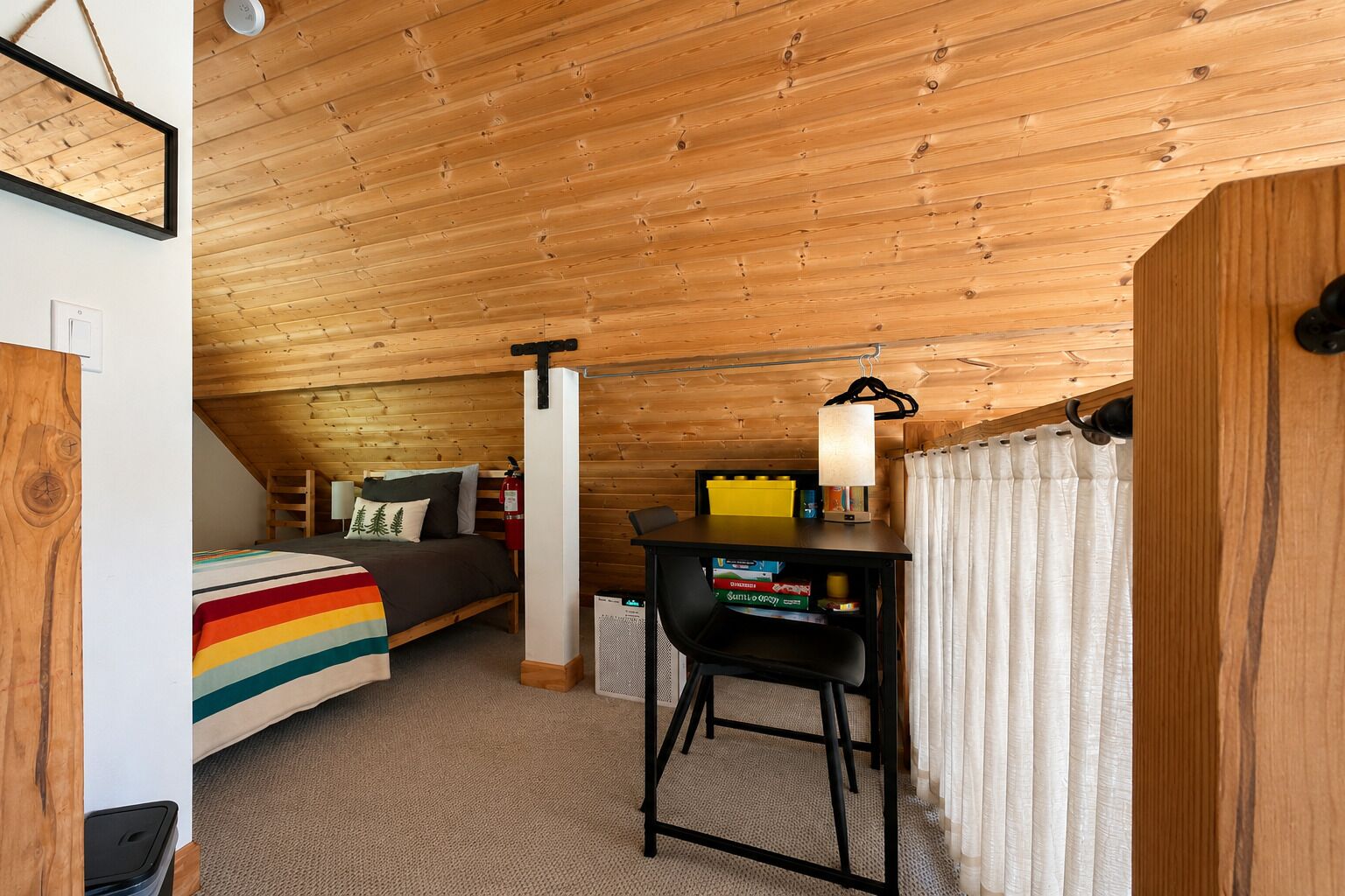 Ein gemütliches Zimmer mit schrägen Wänden, einem Bett mit einer decke in Regenbogenstreifen, einem Schreibtisch mit Büchern und einer Hängelampe.