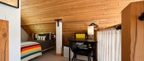Ein gemütliches Zimmer mit schrägen Wänden, einem Bett mit einer decke in Regenbogenstreifen, einem Schreibtisch mit Büchern und einer Hängelampe.