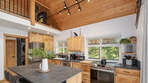 A modern kitchen with wooden cabinets, a central island, stainless steel appliances, and a view of greenery outside. - Raven Haven Lodge 2 Bedroom Home by RedAwning (Leavenworth)
