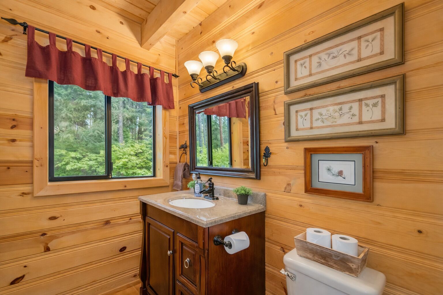 Ein Badezimmer einer Holzhütte mit einem großen Fenster, einem Waschtisch mit Spüle, einem Spiegel und gerahmten Pflanzenbildern an der Wand.