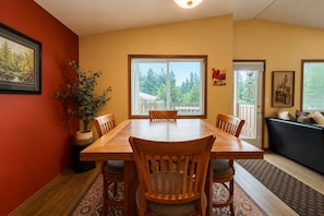 A dining area with a wooden table and chairs, a potted plant, and a view through large windows.