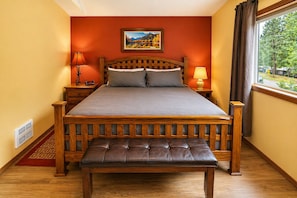 A bedroom with a wooden bed, two bedside tables, a bench, a painting on the wall, and a window with curtains. - Suite Serenity 3 Bedroom Condo by NW Comfy Cabins by RedAwning (Leavenworth)