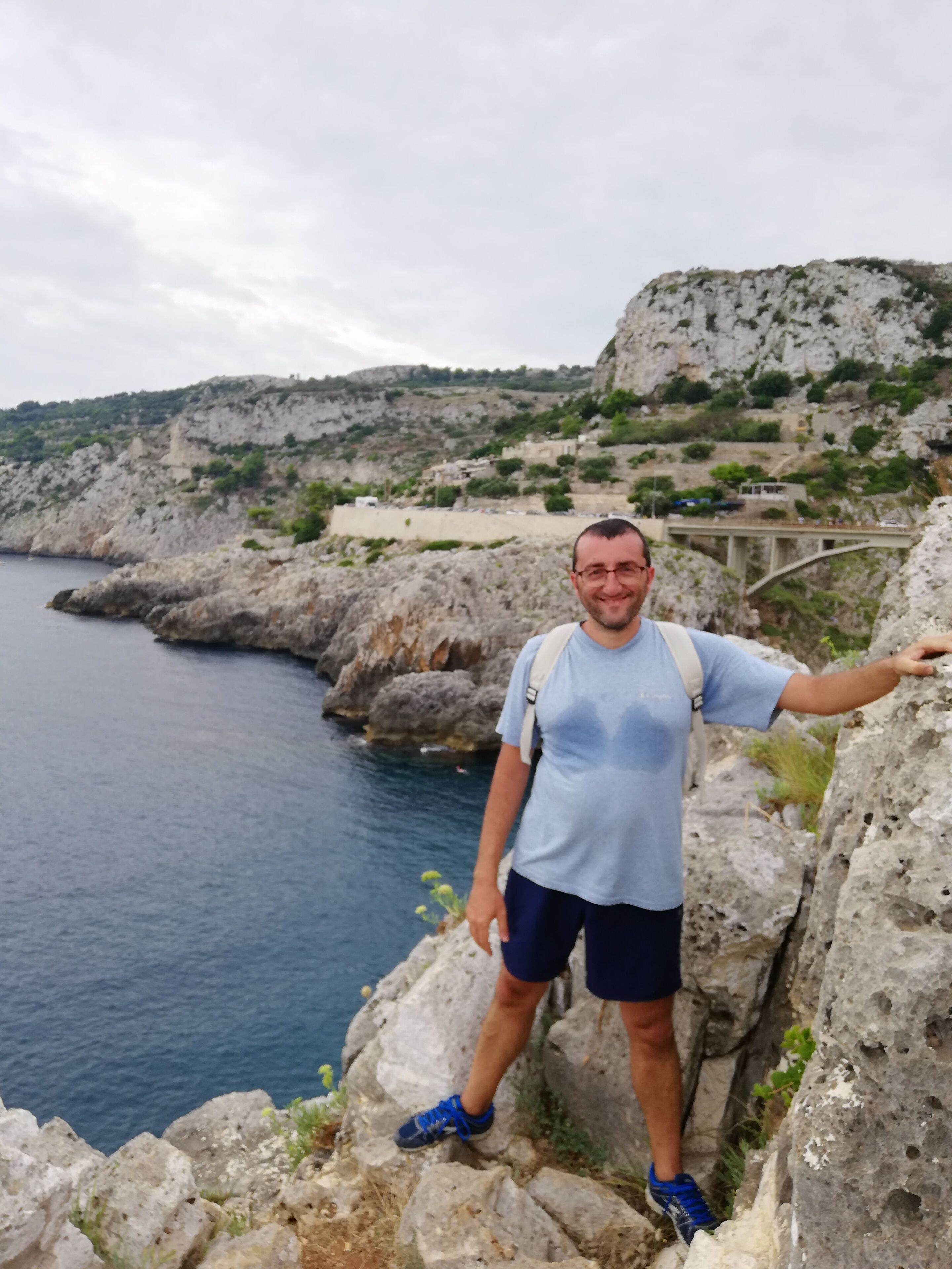 A man standing on a rocky cliff overlooking a body of water, with a bridge and buildings in the background.