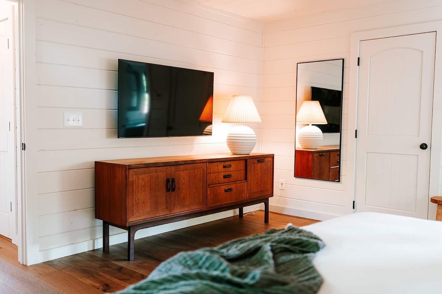 A bedroom with a wooden dresser, a flat-screen TV mounted above it, and a mirror on the wall.