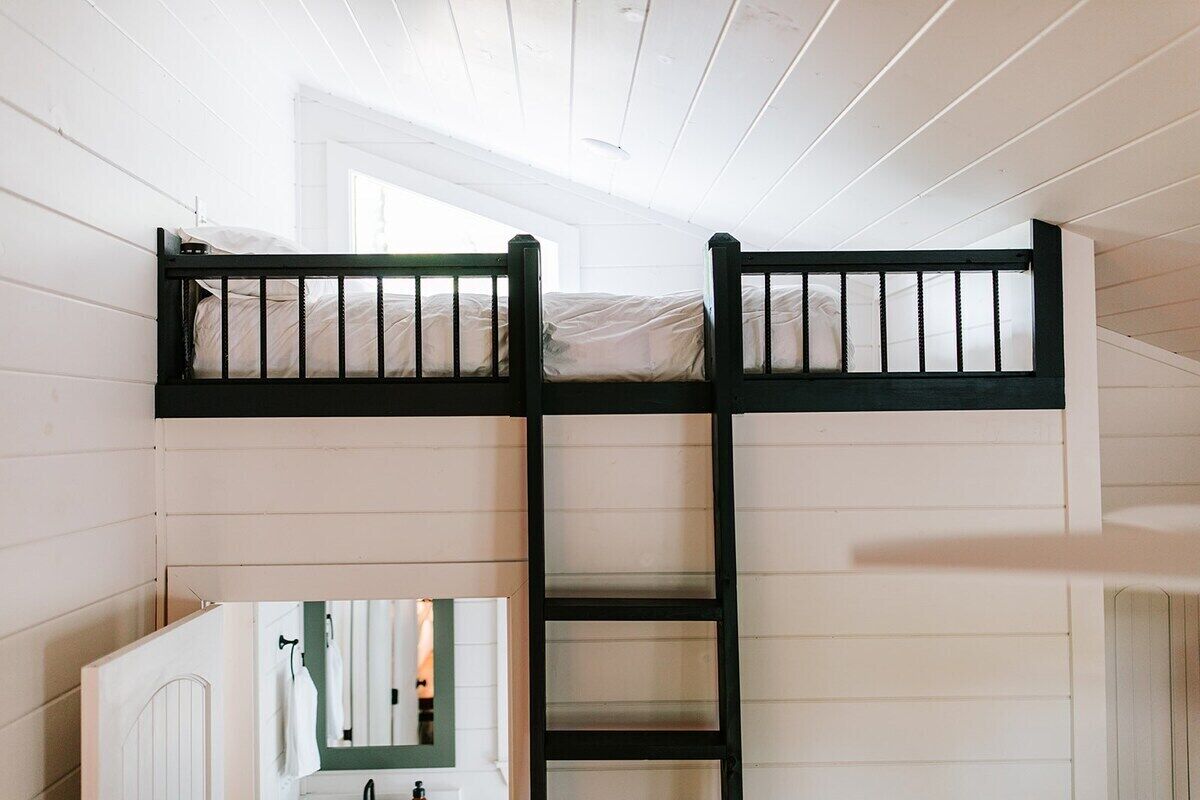 Uma cama beliche com escada, paredes de madeira branca e uma janela.