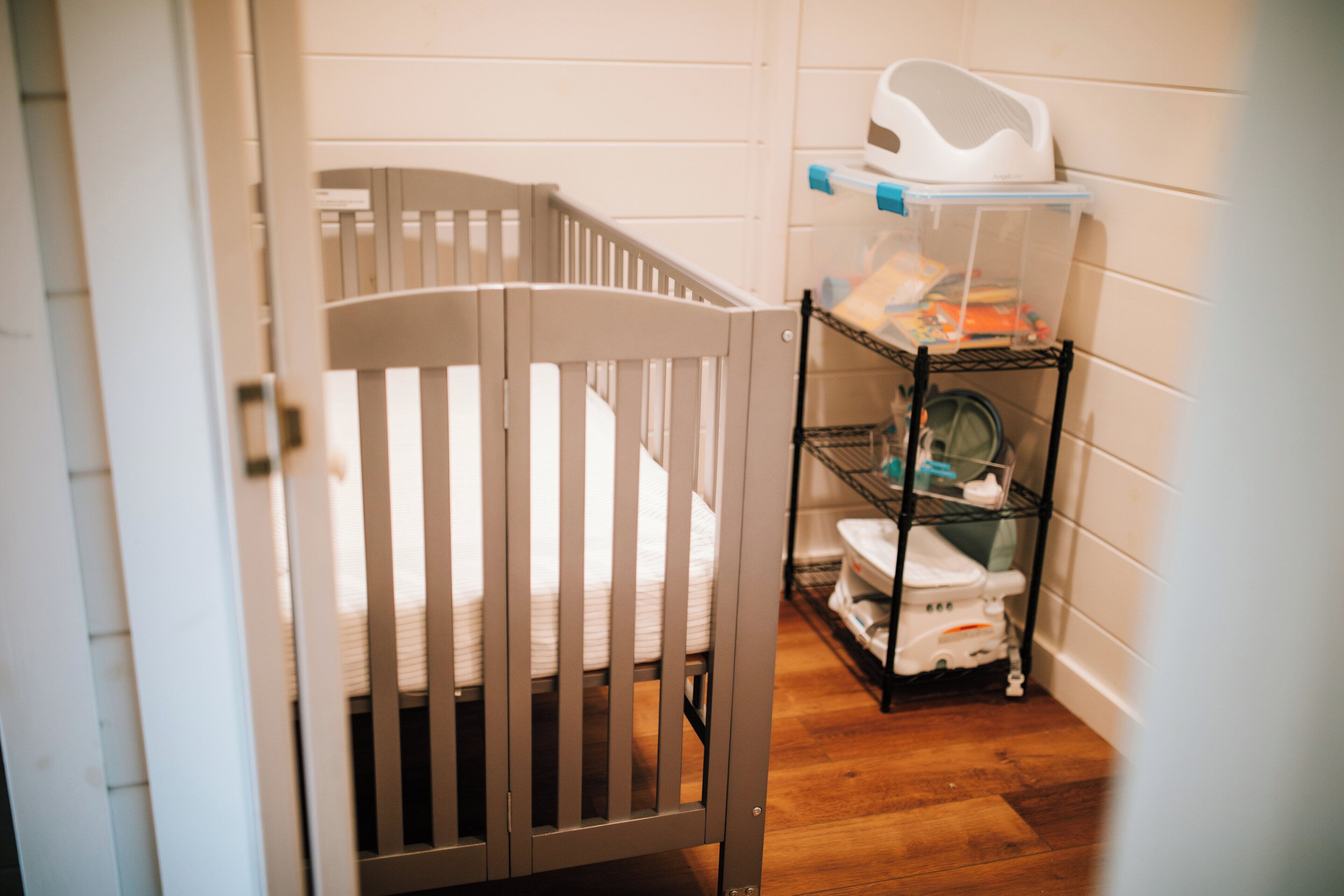 A white crib with a wooden frame, a baby changing station with a white seat and storage shelves, and a white baby walker on a wooden floor.