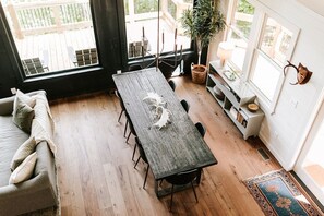 A cozy living room with a wooden dining table, a sofa, and a large window with a view of a wooden deck. - Luxe 4BR Designer Cabin  Hot Tub • Views • Fire (Cherry Log)
