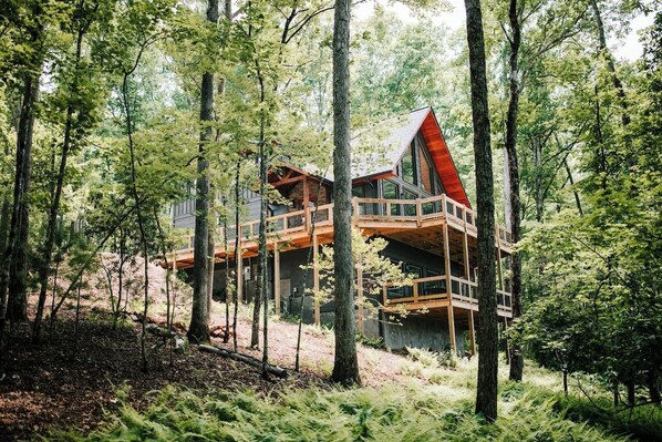 A wooden cabin with a red roof, built on stilts in a forested area. - Luxe 4BR Designer Cabin  Hot Tub • Views • Fire (Cherry Log)