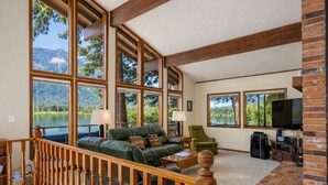 A spacious living room with large windows overlooking a lake, a fireplace, and a cozy seating area with a couch and chairs.