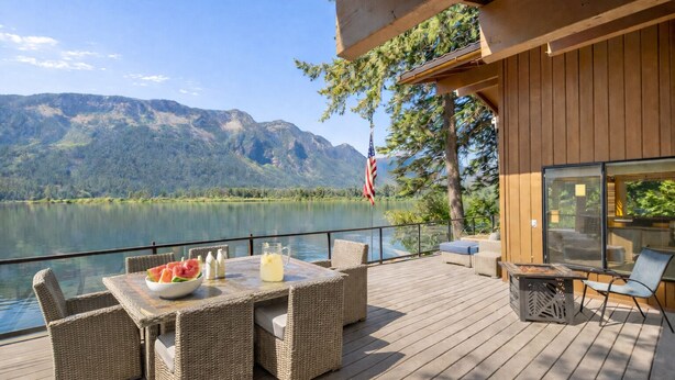 A deck with wicker furniture, a table with a bowl of fruit and drinks, and a view of a lake and mountains.