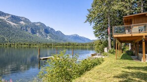 A lakeside cabin with a deck overlooking the water, surrounded by mountains and greenery.