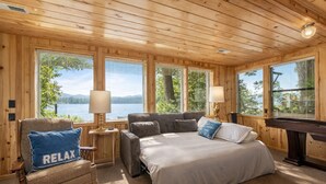A cozy cabin bedroom with a bed, a chair, a desk, and large windows overlooking a lake.