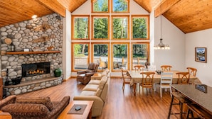 A spacious interior with a stone fireplace, wooden ceiling, and large windows offering a view of the forest. - Getaway Moose 3 Bedroom Home by NW Comfy Cabins by RedAwning (Leavenworth)