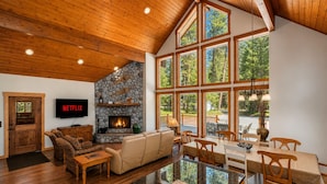 A spacious living room with a stone fireplace, wooden ceiling, and large windows overlooking a forest. - Getaway Moose 3 Bedroom Home by NW Comfy Cabins by RedAwning (Leavenworth)
