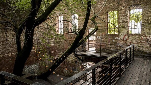An indoor wooden deck with a metal railing, surrounded by brick walls and trees.