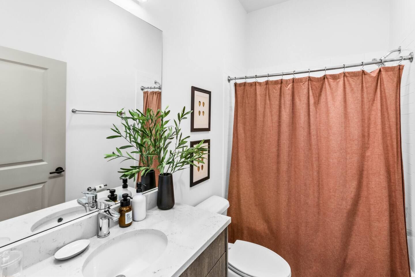 Casa de banho moderna com lavatório branco, espelho e sanita. Tem uma cortina de chuveiro e uma planta num vaso sobre a bancada.