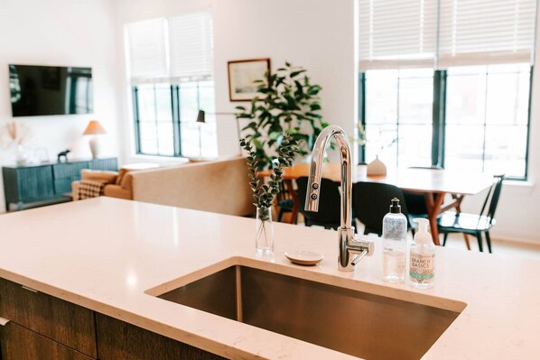 A kitchen with a stainless steel sink, a vase with green leaves, and two bottles of hand soap.