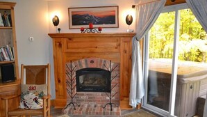 Ein gemütliches Wohnzimmer mit Kamin, Bücherregal, einem Stuhl mit Ski-Motiv und einem großen Fenster mit Blick ins Freie.