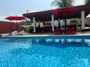 A swimming pool with lounge chairs and umbrellas, a covered seating area, and a white car parked nearby. - Beautiful Villa With Swimming Pool in Assinie (Assinie)