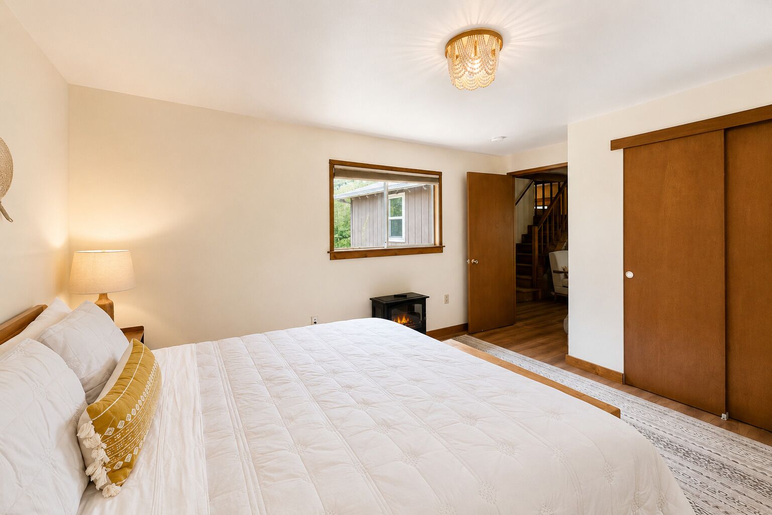 Un dormitorio con una cama grande, una lámpara de noche, una mesita, una ventana y una puerta que da a una escalera.