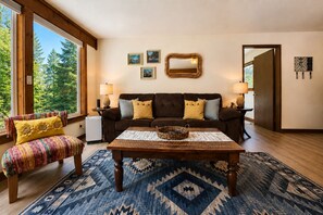 A living room with a sofa, armchairs, a coffee table, and a rug. There are framed pictures on the wall, a mirror, and a door leading outside.