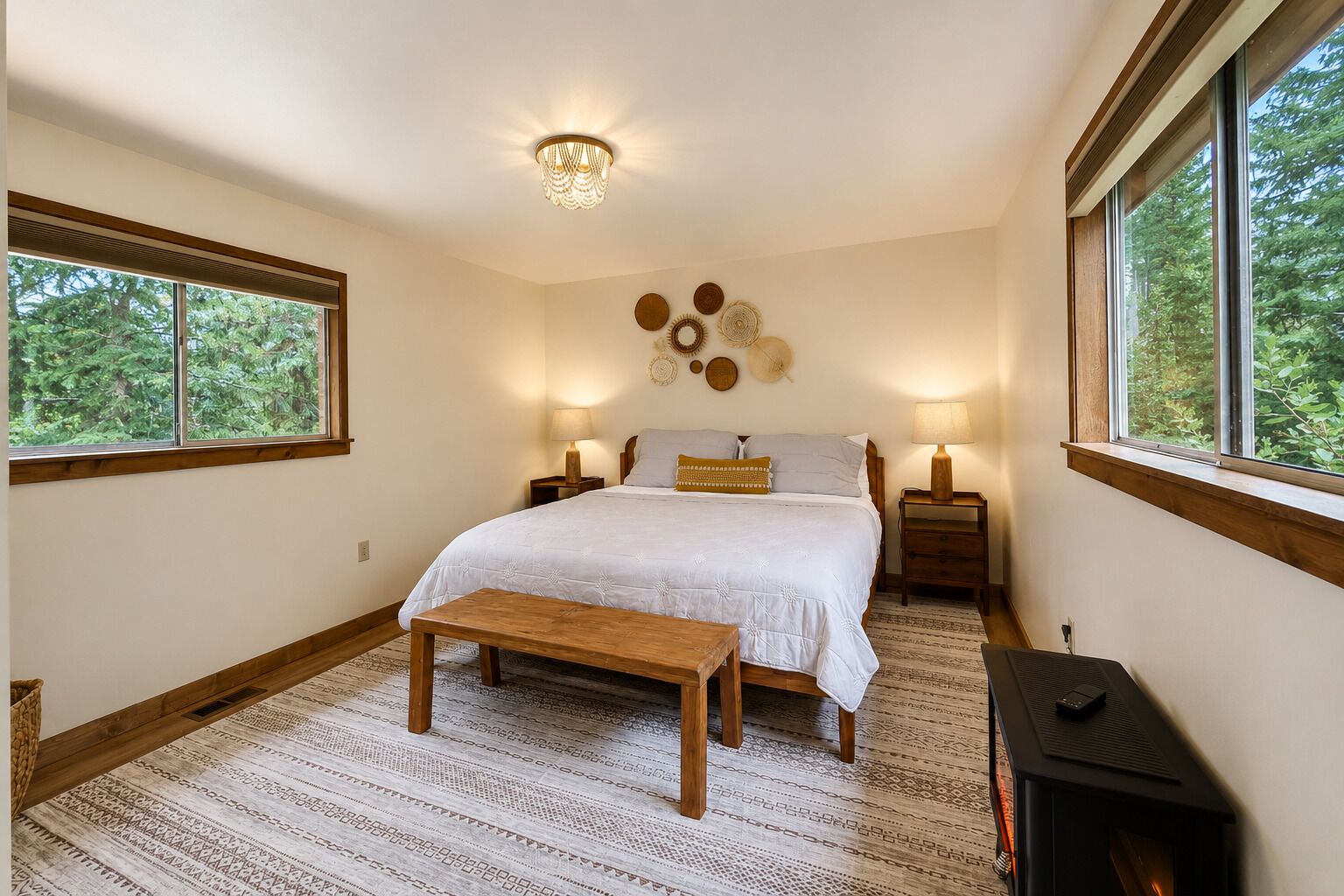 A bedroom with a bed, bedside tables, a bench, a fireplace, and a view of trees through the windows.