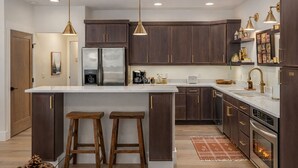 A modern kitchen with dark wood cabinets, stainless steel appliances, and a white marble countertop. There are two wooden stools at the island, and pendant lights hang above. - Bavarian Mountain Suite 2 Bedroom Condo by NW Comfy Cabins by RedAwning (Leavenworth)