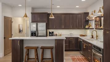 A modern kitchen with dark wood cabinets, stainless steel appliances, and a white marble countertop. There are two wooden stools at the island, and pendant lights hang above.