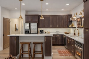 A modern kitchen with dark wood cabinets, stainless steel appliances, and a white marble countertop. There are two wooden stools at the island, and pendant lights hang above.