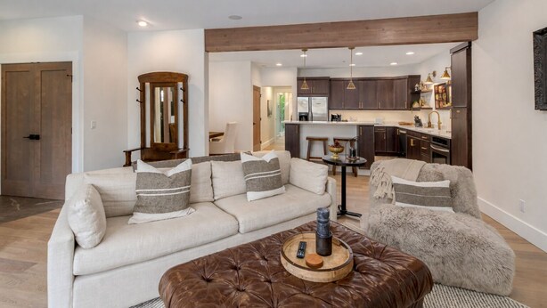 A modern living room with a sofa, armchair, and ottoman, a wooden accent wall, and a kitchen in the background.