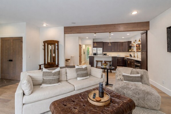 Una sala de estar moderna con un sofá, un sillón y un reposapiés, una pared de acento de madera y una cocina al fondo.