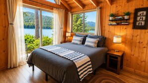 Une chambre avec un grand lit, des murs en bois, une vue sur un lac et une étagère remplie de livres et de plantes.