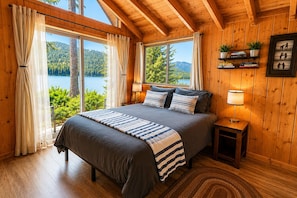 A bedroom with a large bed, wooden walls, a view of a lake, and a shelf with books and plants.