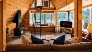 A cozy cabin interior with a loft bedroom, a fireplace, a TV, and a view of a lake. - Doc Roy's Legacy On Fish Lake Home by NW Comfy Cabins by RedAwning (Leavenworth)