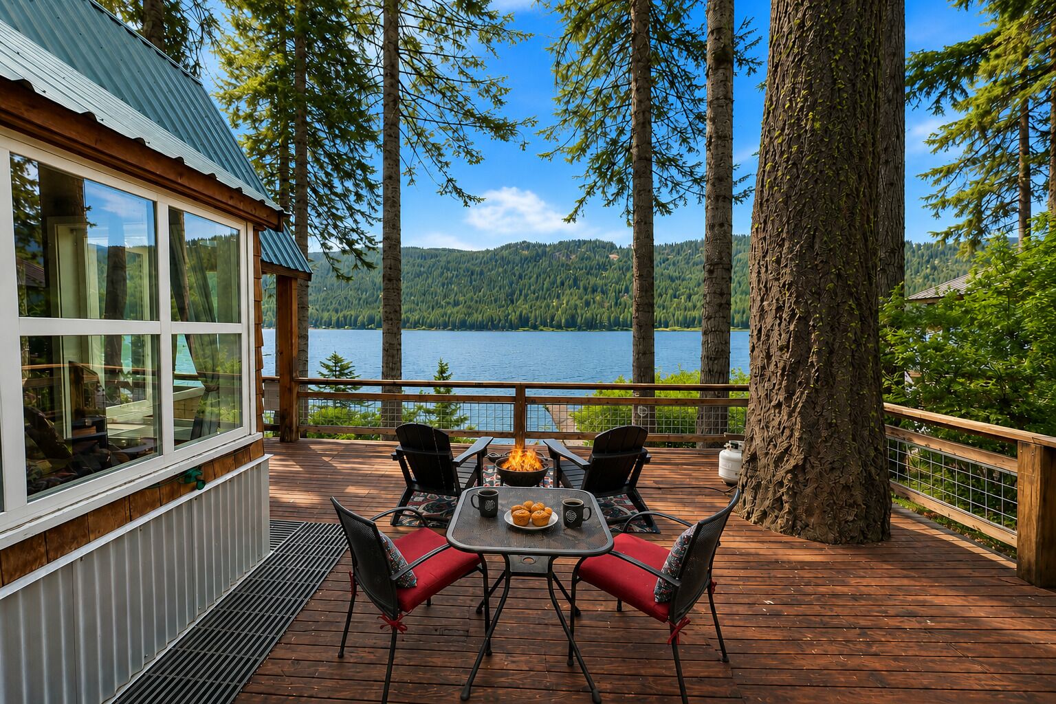 A wooden deck with a table and chairs, overlooking a lake with trees and a cabin.