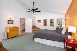 Un dormitorio con cama, cómoda, mesita de noche roja, ventilador de techo y una escalera que lleva a otra habitación.