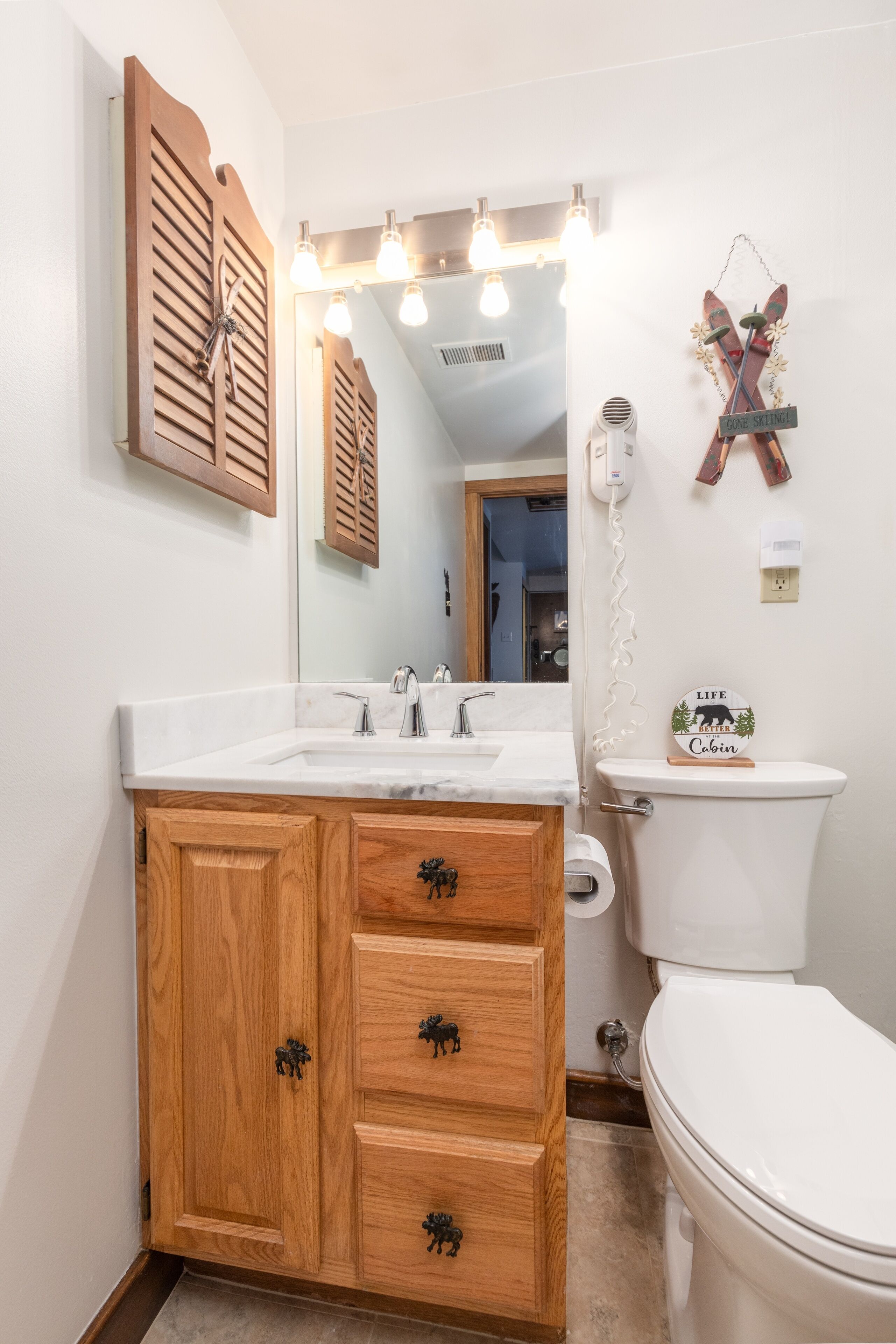 A bathroom with wooden cabinets, a white sink, and a toilet. There are lights above the mirror and a sign on the toilet.