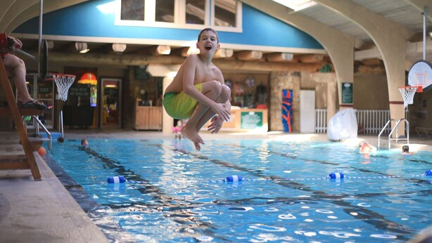 An indoor swimming pool with lanes, a basketball hoop, and a wooden deck area.