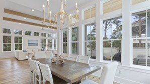 A spacious dining area with a large wooden table and white chairs, large windows offering views of the outdoors, and a chandelier hanging above. - Blue Mountain Beach | Pool | Bikes (Santa Rosa Beach)