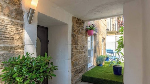 A stone exterior wall, a door, a small window with hanging plants, and a grassy area with chairs.
