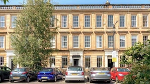 A multi-story building with a row of parked cars in front, a clear sky, and a tree to the left.