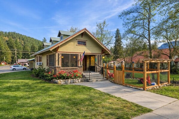A two-story house with a front porch, a small garden with red flowers, and a wooden fence with a gate. - Rockwell Retreat 4 Bedroom Home by NW Comfy Cabins by RedAwning (Leavenworth)