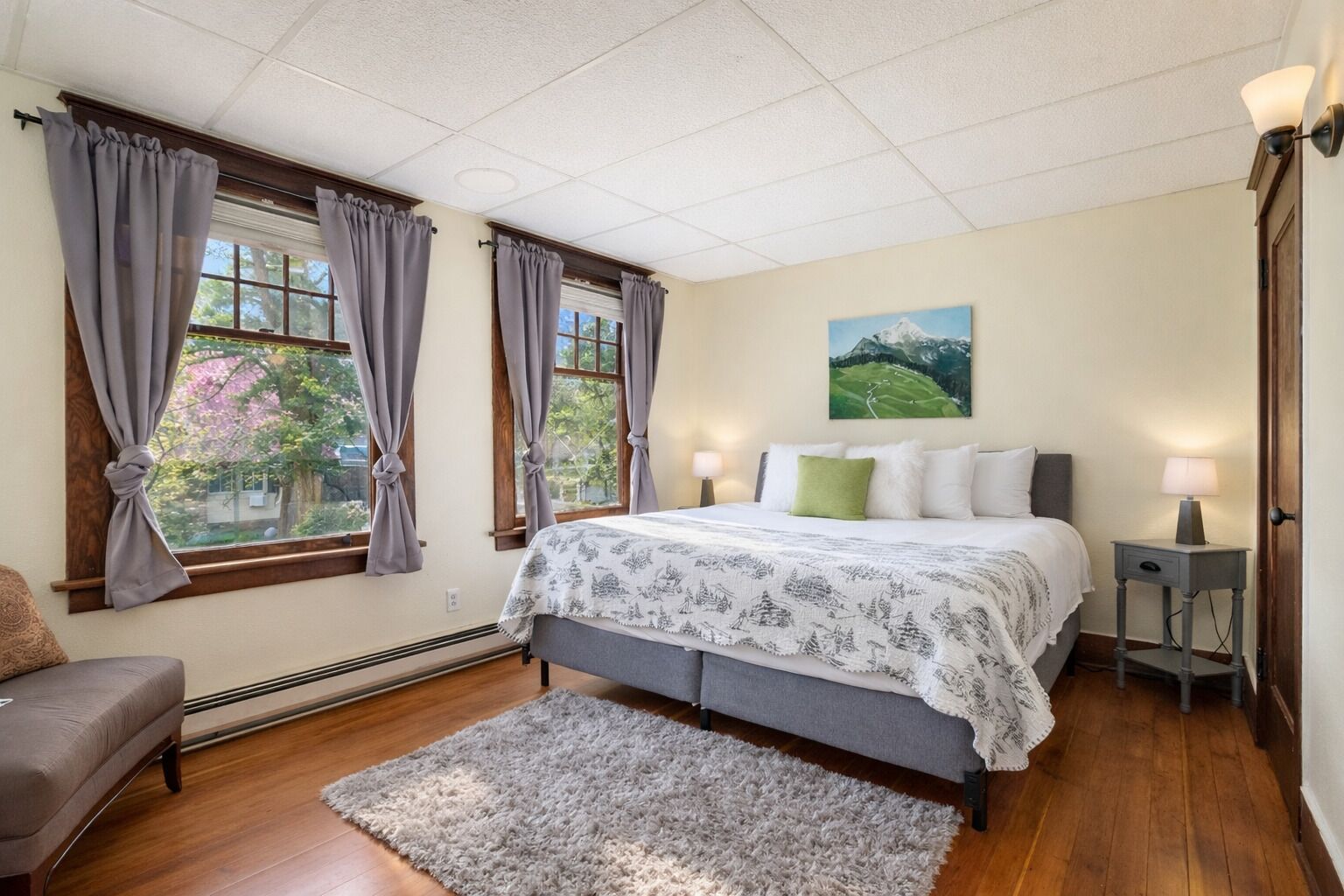 A bedroom with a bed, two bedside tables with lamps, a gray sofa, and a view of a green landscape through the windows.
