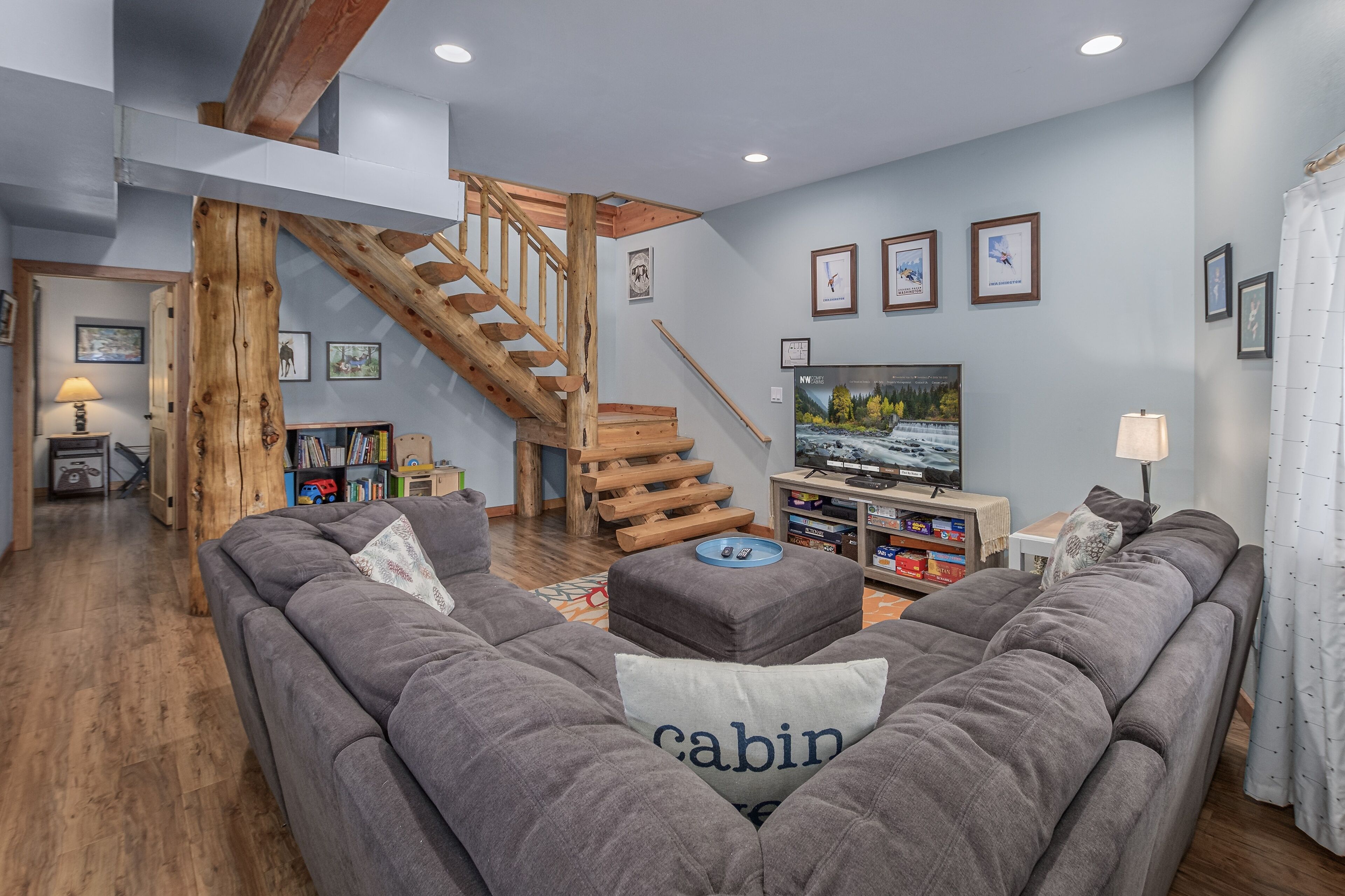 A spacious living room with a grey sofa, wooden staircase, and a television.