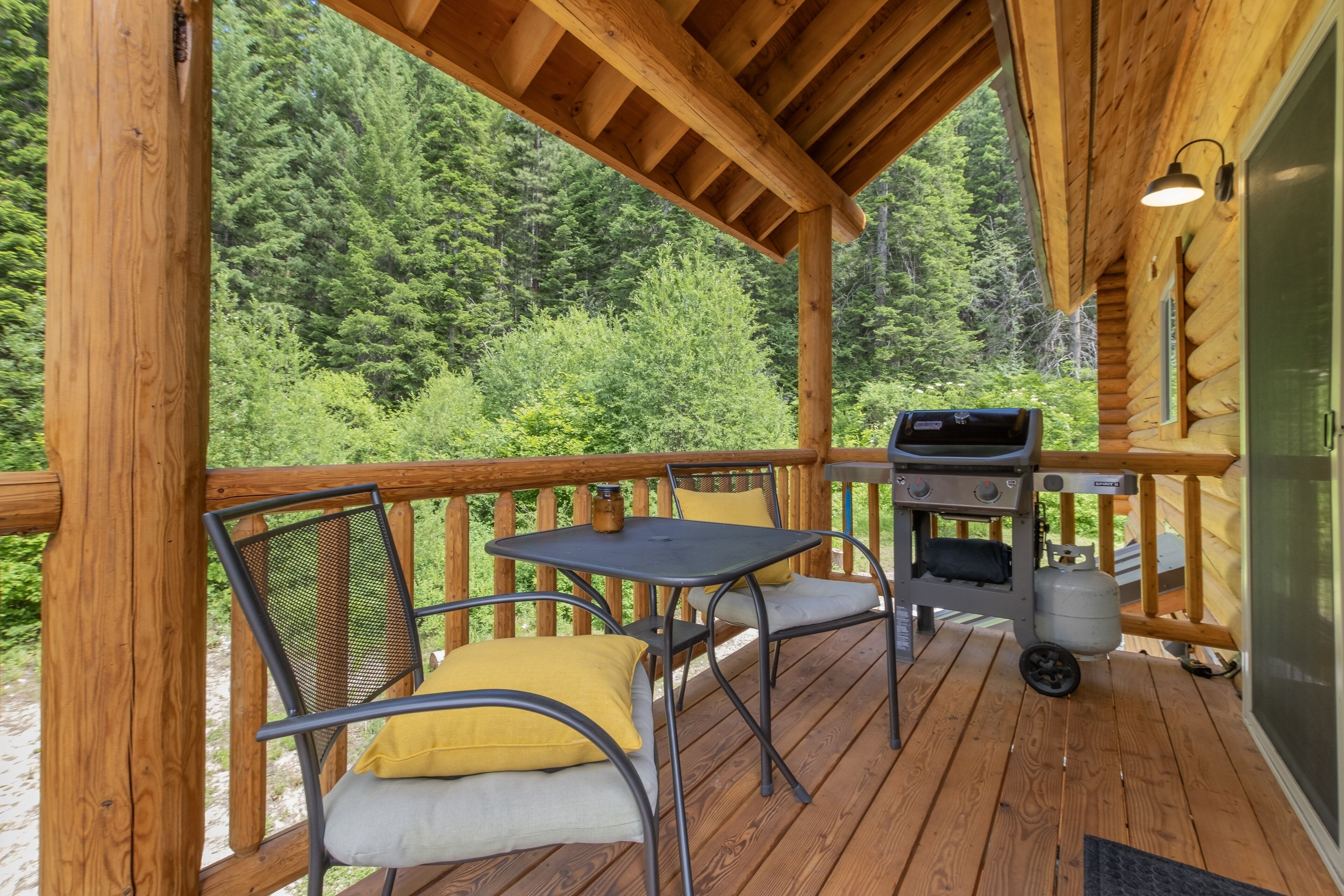 Una terraza de madera con una parrilla y sillas, rodeada de un bosque.