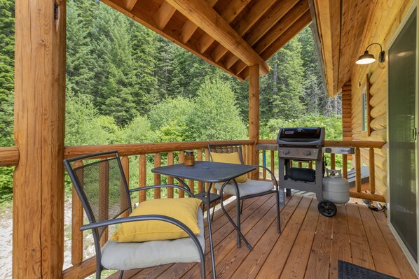 Una terraza de madera con una parrilla y sillas, rodeada de un bosque.