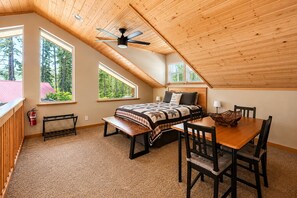 Quarto com cama, escrivaninha e cadeira, ventilador de teto e janelas amplas com vista para árvores.