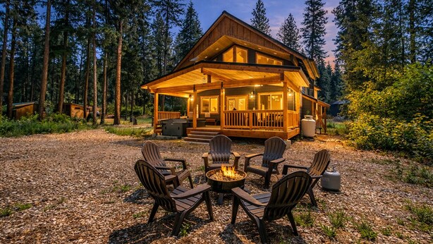 Una cabaña con fachada de madera, una terraza y un pozo de fuego rodeado de sillas Adirondack.