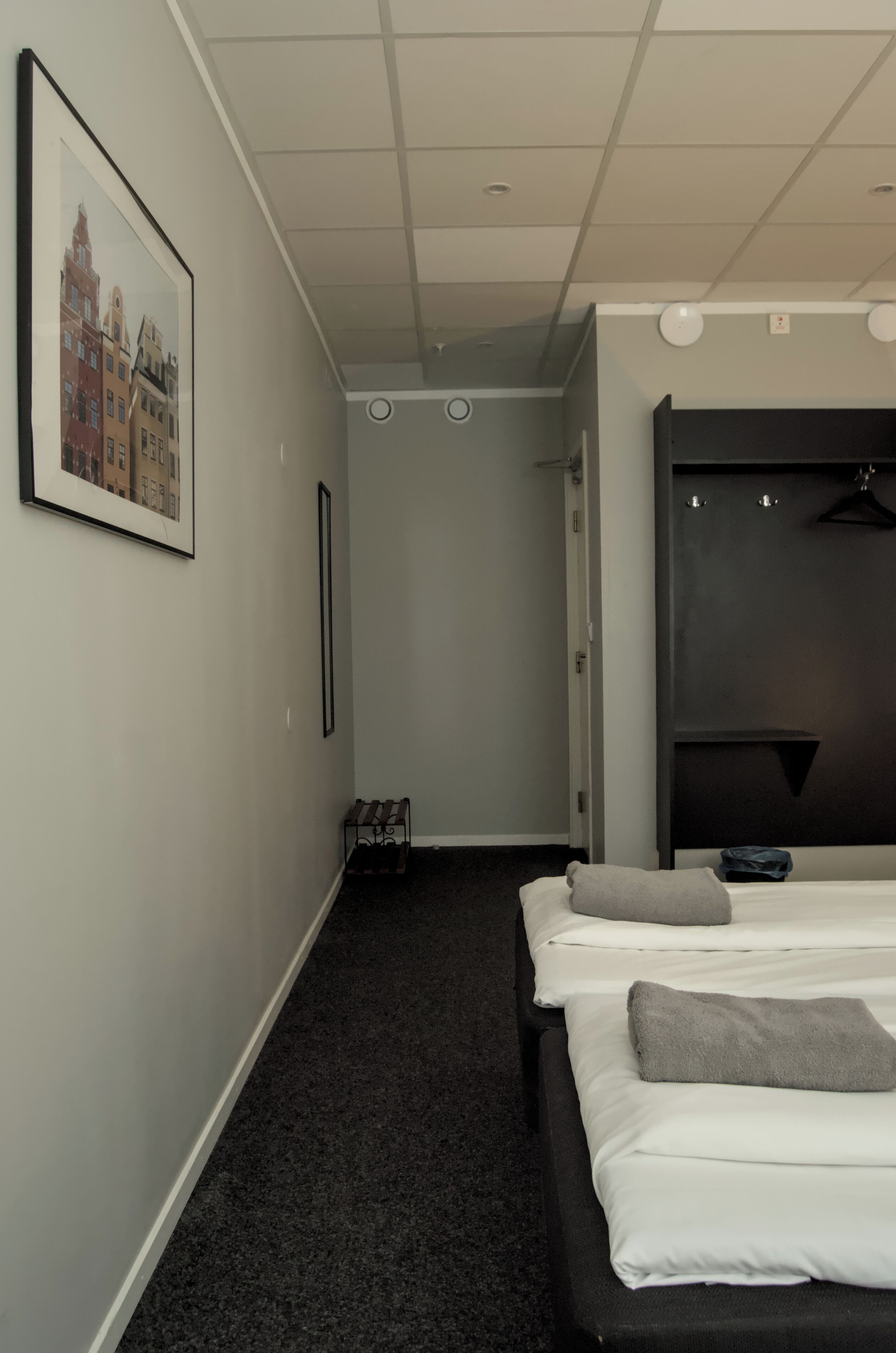 A hotel room with two beds, a framed picture on the wall, a coat rack, and a dark carpeted floor.